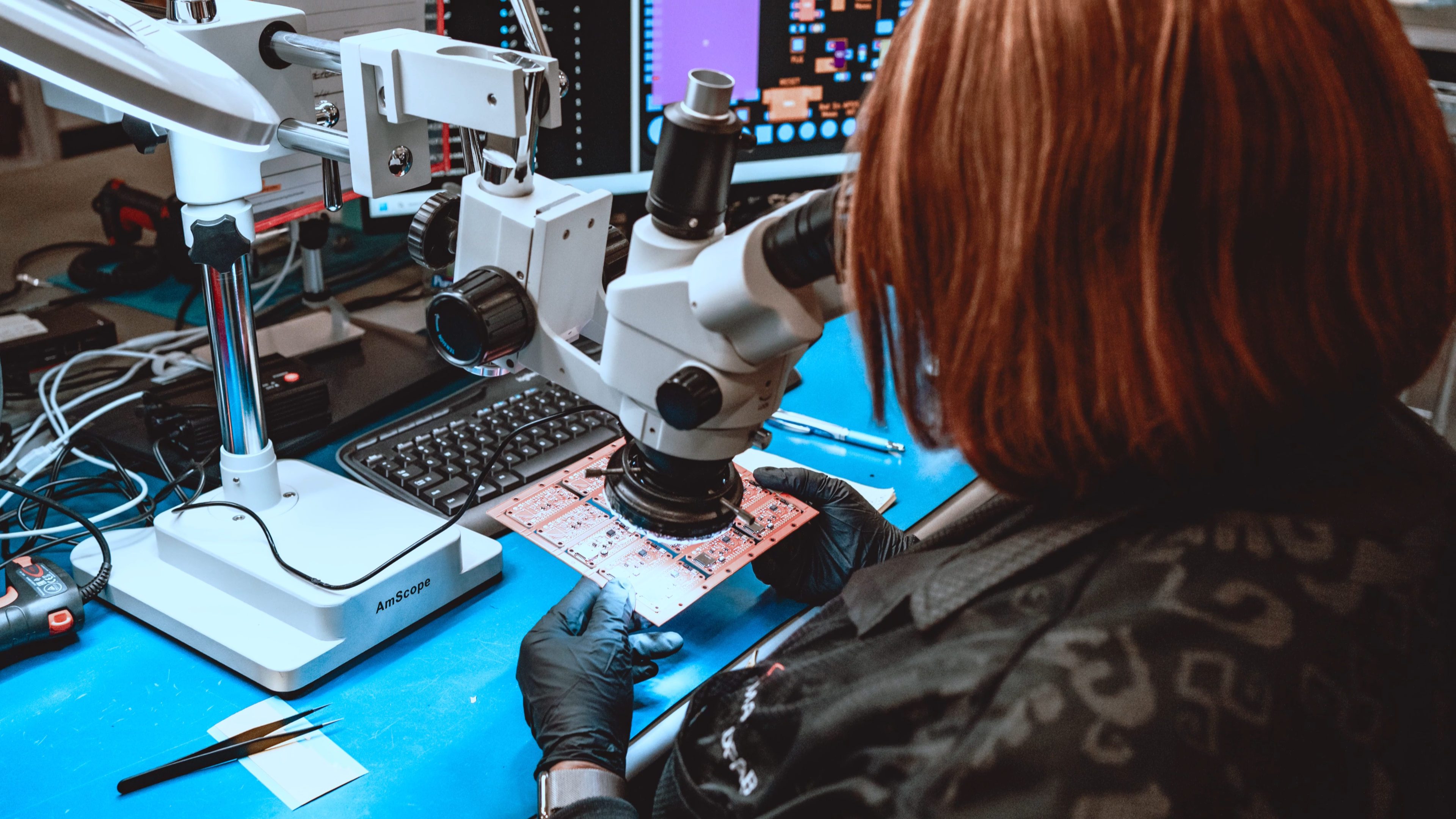 A technician inspecting a panel of assembled Bald SENSE PCBs under a stereo microscope.